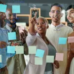 A group of diverse businesspeople are engaged in a lively brainstorming session in a modern office. Gathered around a large glass wall, they use colorful adhesive notes to capture and organize their ideas. Dressed in professional attire, their animated expressions and gestures reflect enthusiasm and collaboration.