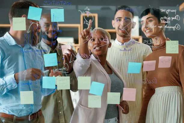 A group of diverse businesspeople are engaged in a lively brainstorming session in a modern office. Gathered around a large glass wall, they use colorful adhesive notes to capture and organize their ideas. Dressed in professional attire, their animated expressions and gestures reflect enthusiasm and collaboration.