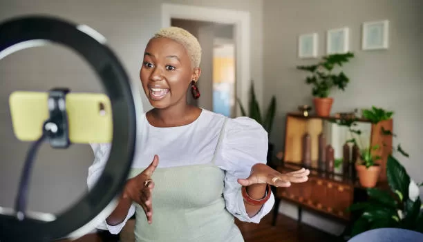 Smiling young African female influencer standing in her living room at home and talking during a vlog post using a smart phone